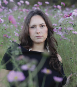 A woman with long brown hair wearing a black off-the-shoulder top stands in a field of pink and purple flowers, looking directly at the camera with a neutral expression.