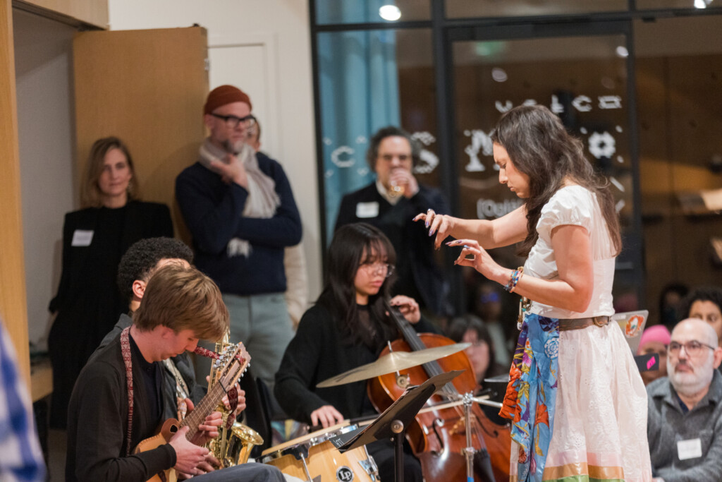 A woman conducts a small group of musicians playing various instruments indoors, while an attentive audience watches in the background.