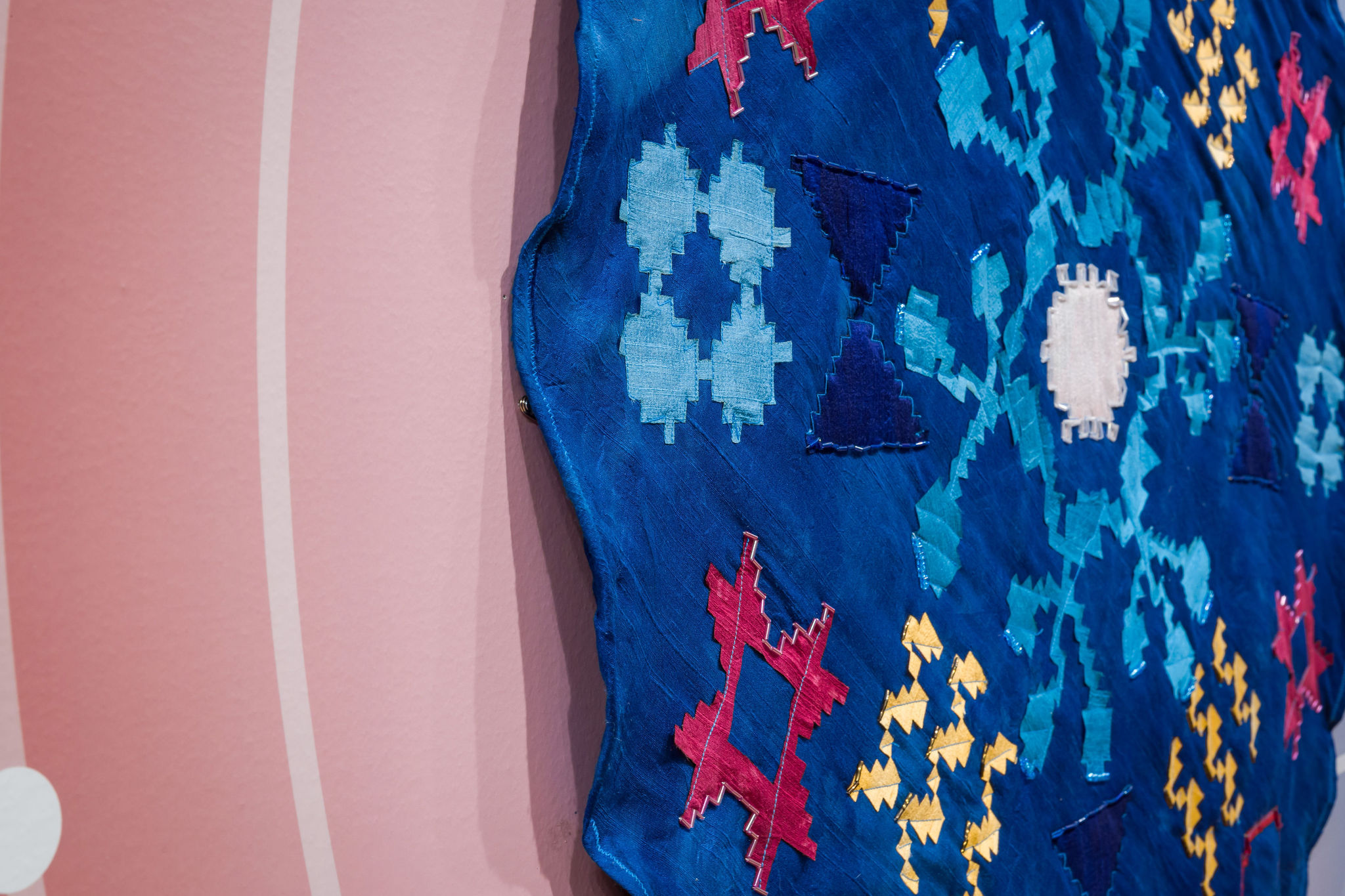 A close-up of a blue textile with geometric patterns in red, yellow, light blue, and white, displayed against a pink wall with white curved lines.