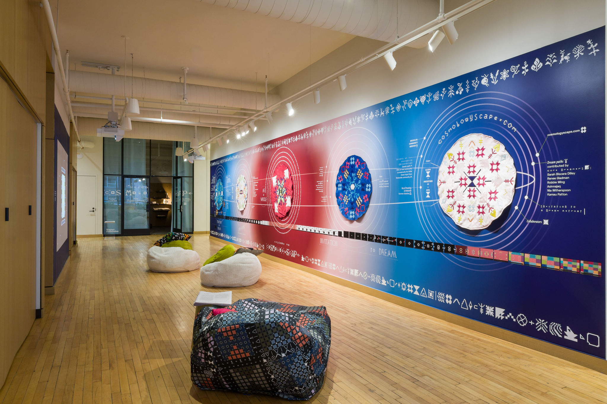 A modern gallery space with wooden floors, colorful bean bag chairs, and a large, vibrant mural featuring scientific diagrams, patterns, and text along one wall. Track lighting illuminates the artwork.
