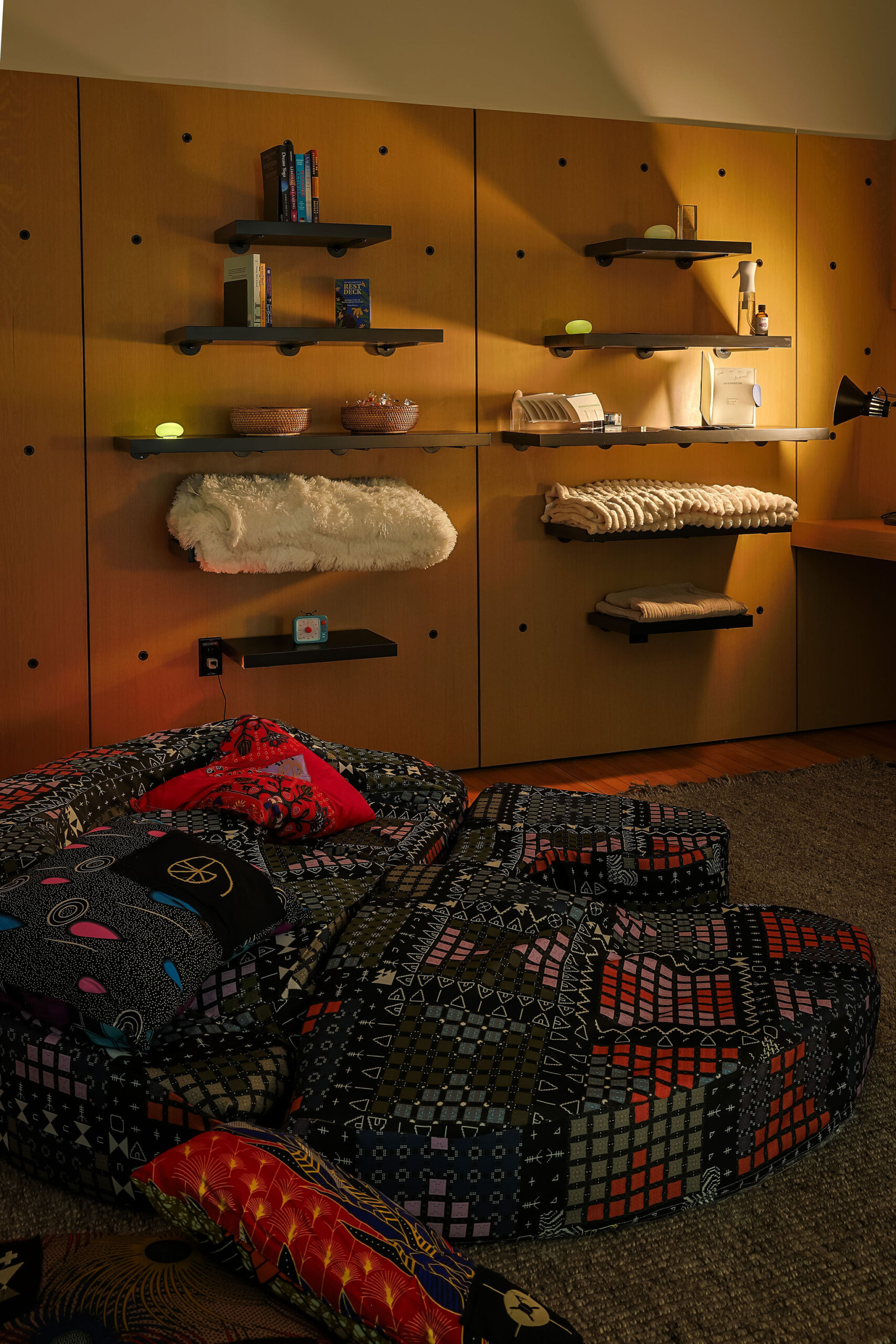 A cozy room with a patterned sofa, colorful pillows, a wooden wall featuring floating shelves holding books, decor, and cushions, and a soft carpet on the floor. Warm ambient lighting creates a relaxed atmosphere.