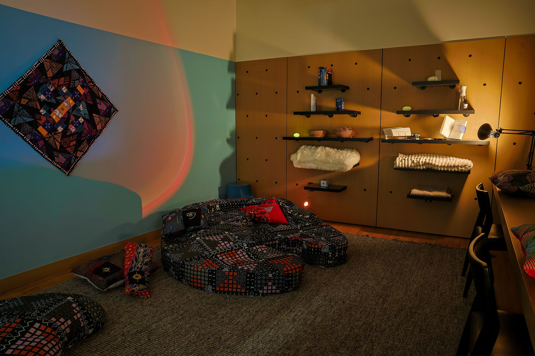 A cozy room with patterned floor cushions, wall-mounted shelves holding various items, a colorful textile art piece on a blue wall, ambient lighting, and a desk with chairs on a gray carpet.