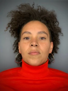A person with curly dark hair, wearing large hoop earrings and a bright red turtleneck, looks directly at the camera against a plain gray background.