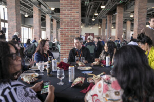 A group of people sit around a round table with food, drinks, and snacks, having a conversation in a large indoor space with brick pillars and windows. Some are eating, chatting, or using their phones.