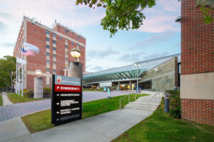 A hospital entrance with a sign for emergency, main entrance, and parking. The building has brick and glass sections, with an American flag, trees, and a sidewalk leading to the door.