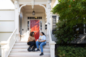 Two people sit on the front steps of a house with a bright red door, smiling and talking. They appear relaxed and happy, enjoying a conversation on a sunny day.
