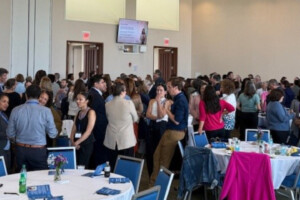 A large group of people are mingling and conversing in a bright conference room with round tables covered in white tablecloths, blue chairs, and informational materials. A digital display screen hangs on the wall.