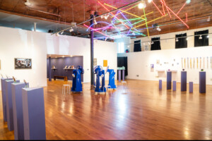 A contemporary art gallery with wooden floors, white walls, colorful neon lights on the ceiling, blue interactive machines, display pedestals, and various artworks showcased around the room.
