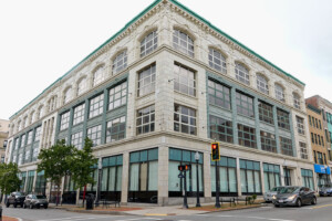 A large, historic, four-story building with many windows on a city street corner, featuring ornate white stonework on the upper floors and greenish-blue accents on the lower facade. Cars and a traffic light are in front.