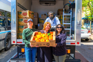 Four people stand behind an open truck filled with produce boxes, smiling and holding a crate of yellow and orange bell peppers. The scene is outdoors, and a bus is visible on the left.