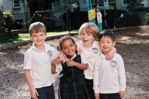 Four young children smile and pose together outdoors at a playground, wearing school uniforms. Three boys wear white polos with a school logo, and one girl in a plaid dress is hugged by the others. Trees and playground equipment are in the background.