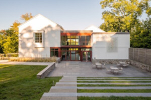 Modern two-story building with white exterior, large glass entrance, and patio area with tables and chairs. Surrounded by trees and grass, with stone steps leading up to the patio.