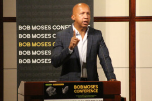 A man in a checkered suit speaks at a podium with a sign reading Bob Moses Conference. He gestures with one hand raised, and a large conference banner is displayed behind him.