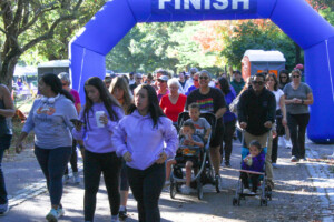 A diverse group of people, including families with strollers, are walking under a blue inflatable finish line at an outdoor event on a sunny day. Trees and portable restrooms are visible in the background.