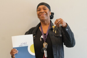 A smiling woman wearing a black headscarf holds up a set of keys in one hand and a Way Finders folder in the other, standing against a plain light-colored background.