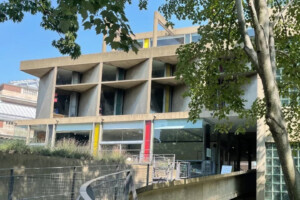 A modern concrete building with geometric design, large windows, colorful vertical panels, and surrounded by trees. The image shows a sloped walkway leading to the entrance.