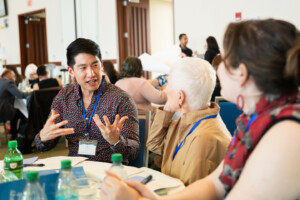 A man wearing a conference badge gestures while speaking to two people at a round table during a meeting or workshop. Other attendees are seen in the background. Bottles and papers are on the table.