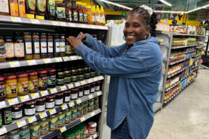 A smiling woman in a blue denim shirt stocks jars on a grocery store shelf, surrounded by various sauces and spreads in a brightly lit aisle.