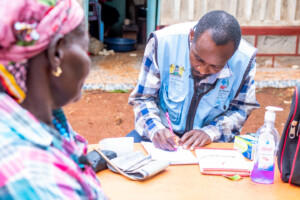 A healthcare worker in a blue vest writes in a notebook at a table outdoors while a woman in colorful clothing sits across from him. Medical supplies, including sanitizer and a blood pressure monitor, are on the table.