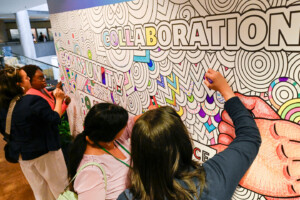 Four people color a large, intricate mural with the words “COLLABORATION” and “COMMUNITY” outlined in bold letters. The mural features abstract patterns and designs, partially filled in with vibrant colors.