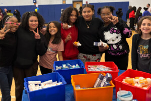 A group of six smiling children stand together in a gymnasium behind a table with red, orange, blue, and yellow bins filled with toiletries and hygiene products. Some kids hold up peace signs and others hold items.