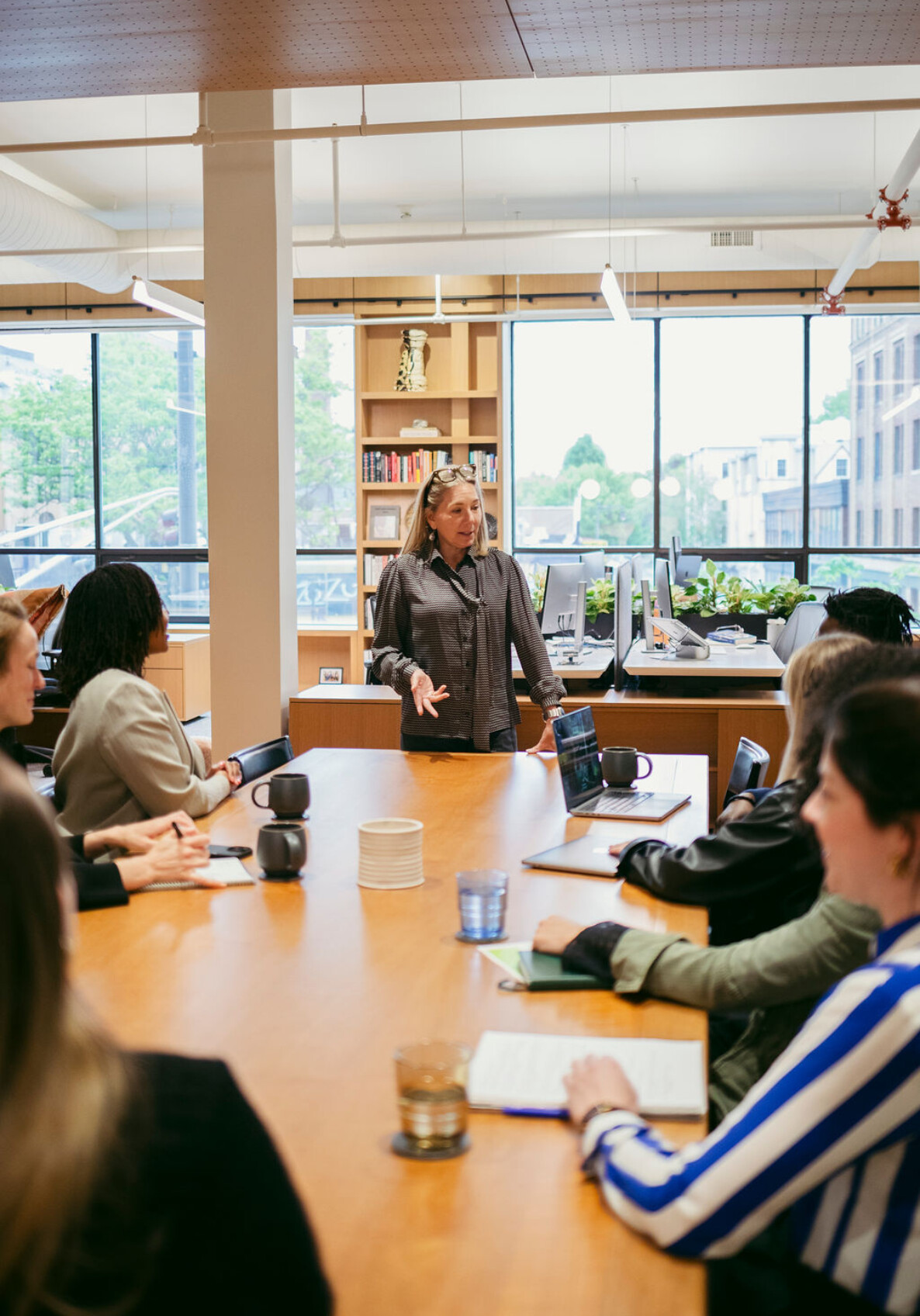 A woman stands at the head of a conference table, speaking to a group of people seated around her in a modern office with large windows and natural light.