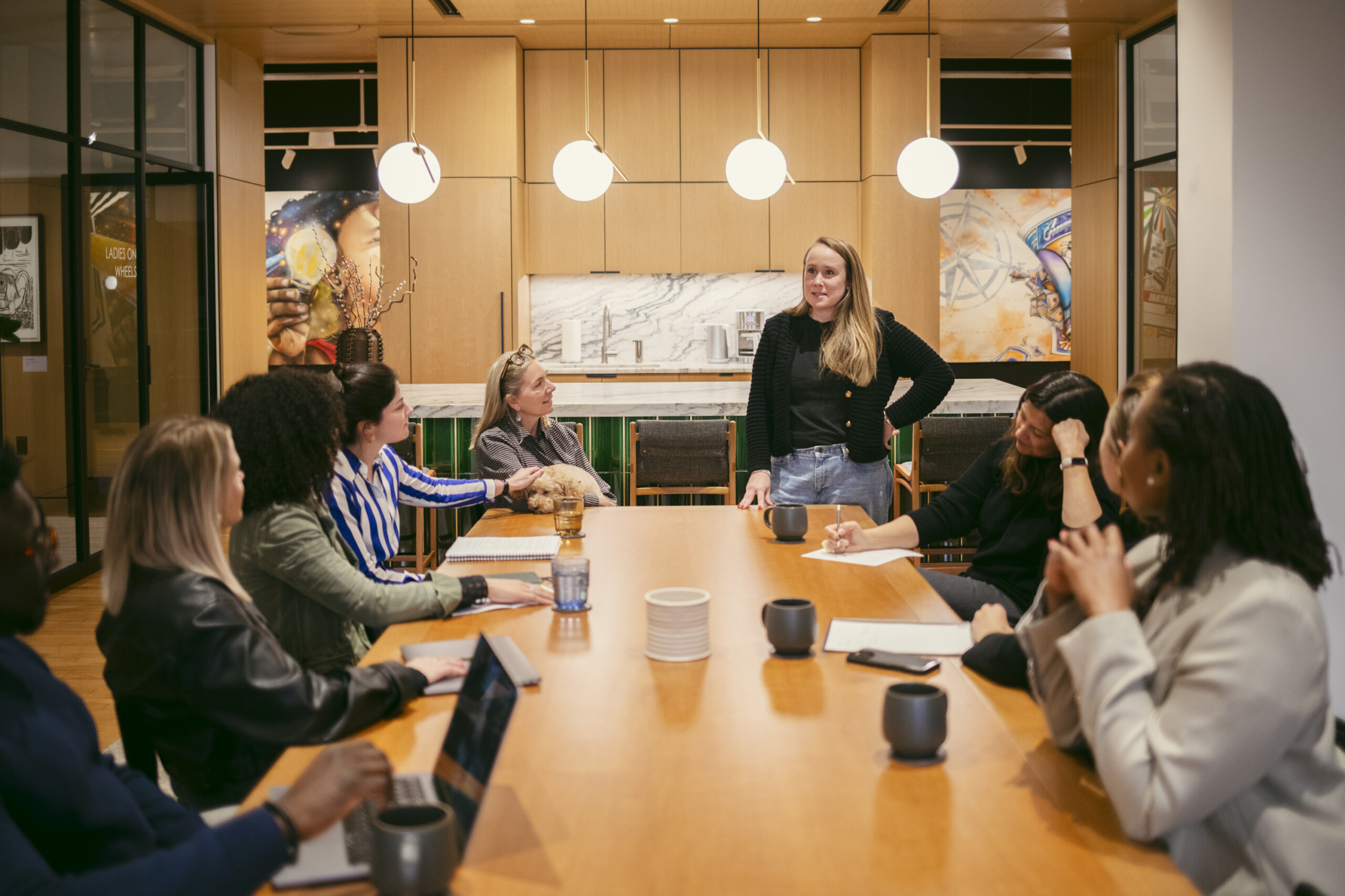 A woman stands and speaks to a group of colleagues seated around a conference table in a modern office, with coffee mugs, laptops, and artwork visible in the background.
