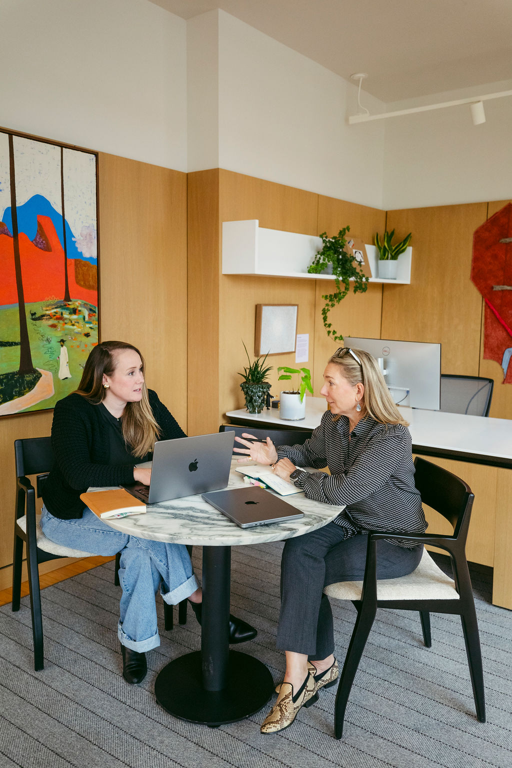 Two women sit at a round marble table in a modern office, having a discussion with laptops open in front of them. The office has wood panel walls, colorful artwork, and plants on shelves.