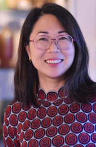A woman with long dark hair and glasses smiles at the camera. She is wearing a patterned sweater with red and blue circular designs. The background is softly blurred.