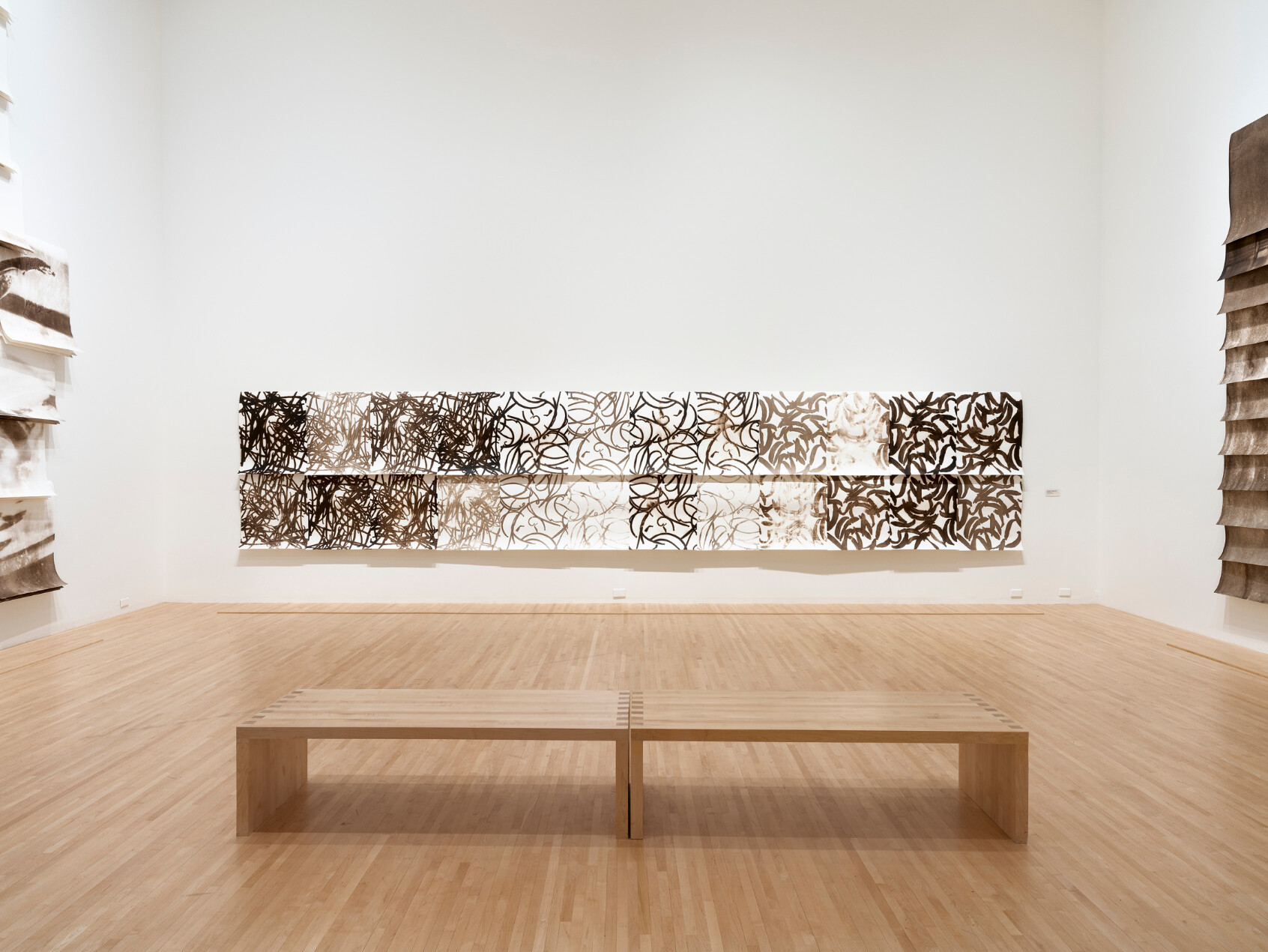 A minimalist art gallery with light wooden floors, white walls, a simple wooden bench, and a large abstract wall sculpture featuring intricate, organic cut-out patterns in various shades of brown and white.