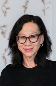 A woman with shoulder-length dark hair and red cat-eye glasses smiles softly while wearing a black top and silver hoop earrings, standing in front of a light background with subtle decorations.