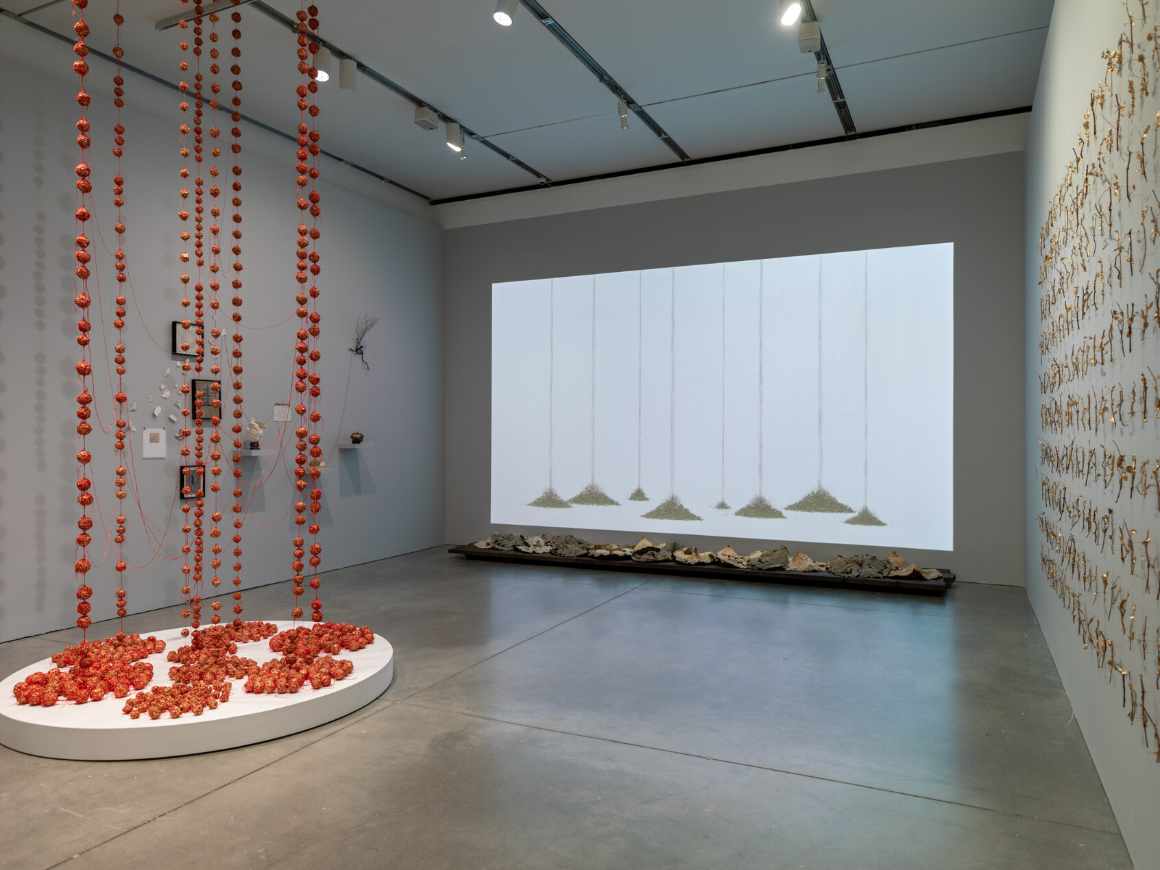 A contemporary art gallery features strings of red objects hanging from the ceiling, a round platform covered in red items, wall-mounted mixed media art, a video projection with sand pendulums, and objects displayed on the floor.