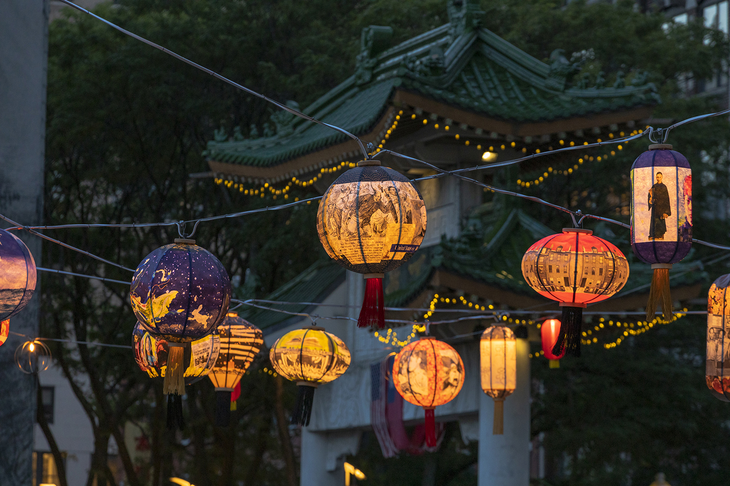 Colorful lanterns with intricate designs hang on strings, illuminated against a backdrop of a traditional green-roofed Asian pavilion and trees, creating a festive atmosphere in the evening.