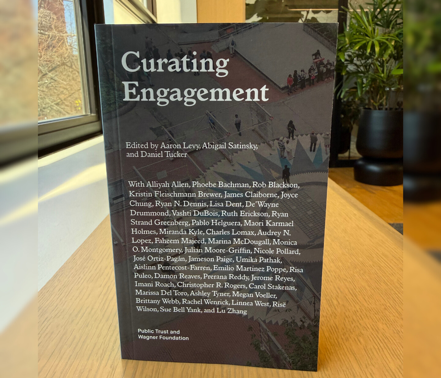 The book Curating Engagement stands upright on a wooden surface, with trees and sunlight visible through a window in the background. The cover lists the editors and contributors in white text on a dark background.