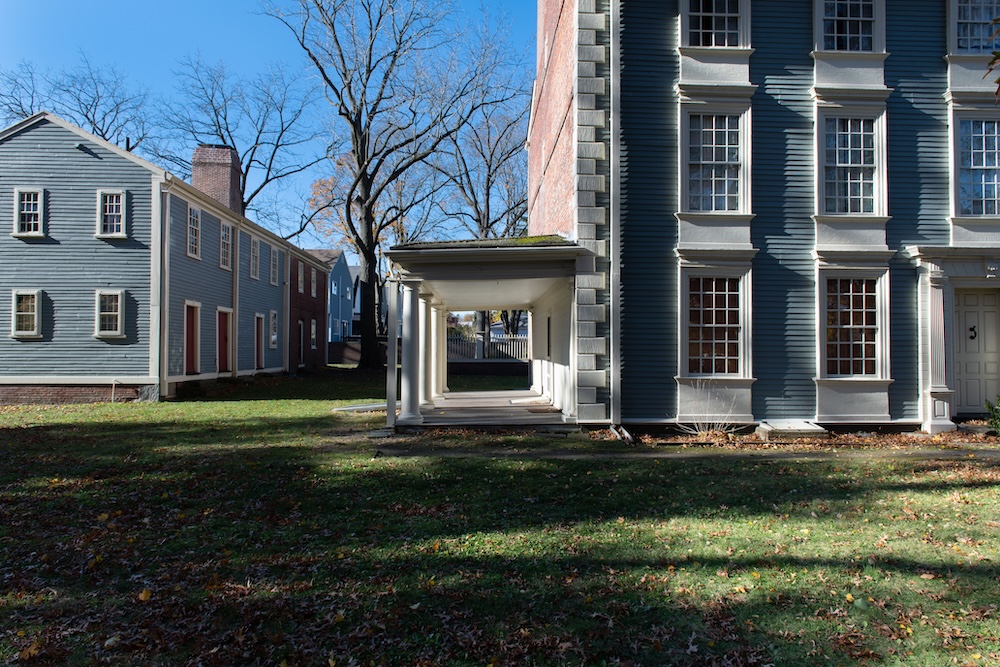 A grassy yard with scattered leaves sits between two historic blue-gray wooden buildings connected by a covered walkway; leafless trees and shadows suggest late autumn.