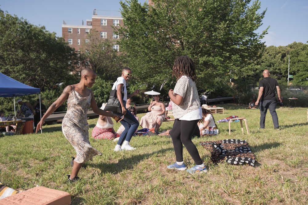 People dance and relax on a grassy lawn during a sunny day. Some are sitting on blankets or low benches, while others stand or dance. There are trees and apartment buildings in the background.