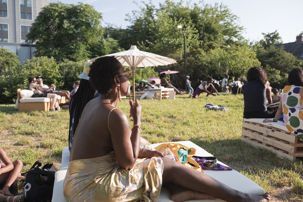 A woman in a yellow dress sits on a white bench outdoors, holding a parasol for shade. People relax on benches around her on a grassy lawn, surrounded by trees and sunlight.