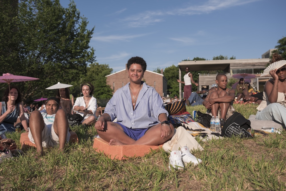 A group of people sit on the grass at an outdoor event on a sunny day. A smiling person in a blue shirt sits cross-legged in the foreground, surrounded by others relaxing and chatting. Trees and buildings are in the background.