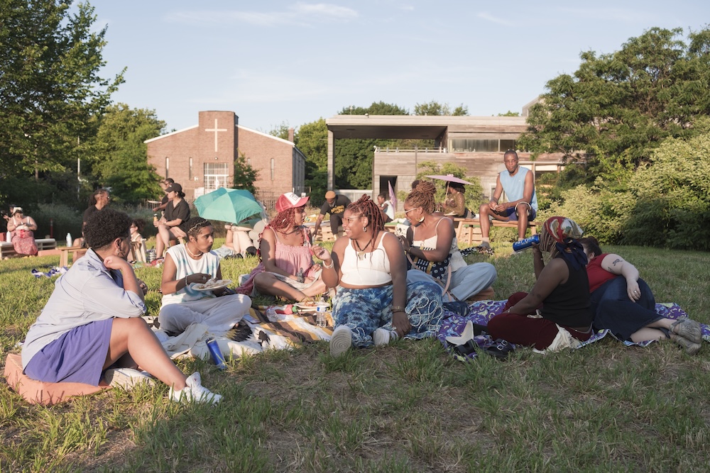 A group of people sit on blankets and grass in a park, enjoying food and conversation on a sunny day. There are trees, buildings, and a church with a cross in the background.