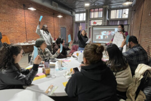 A group of people sit around tables in a brick-walled room, holding up colored cards during a discussion. Two presenters stand at the front, one holding a poster and the other gesturing, with a slideshow on a screen behind them.