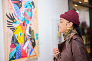 A woman in a red beanie and brown jacket smiles while looking at a colorful, abstract portrait painting in an art gallery. She holds a drink and appears to be enjoying the artwork.