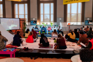 A diverse group of people sits on the floor and cushions, attentively watching a panel of speakers in a bright, modern room with large windows, posters, plants, and a projector screen showing an outdoor photo.