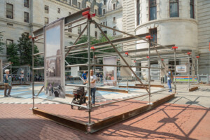 People walk around and examine an outdoor art installation made of metal scaffolding and large photo panels, set in a courtyard surrounded by historic stone buildings. Some people push strollers and observe the displays.