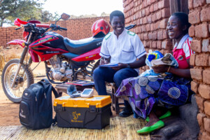 A smiling healthcare worker in uniform sits outside with a woman holding a baby, surrounded by medical supplies, a backpack, and a motorcycle, suggesting a community healthcare visit in a rural area.