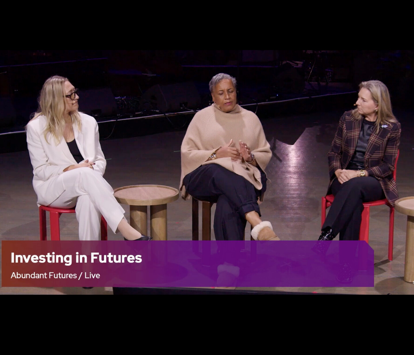 Three women sit on stage having a discussion. They are seated on red chairs with small round tables between them. A purple banner at the bottom reads, “Investing in Futures. Abundant Futures / Live.”.