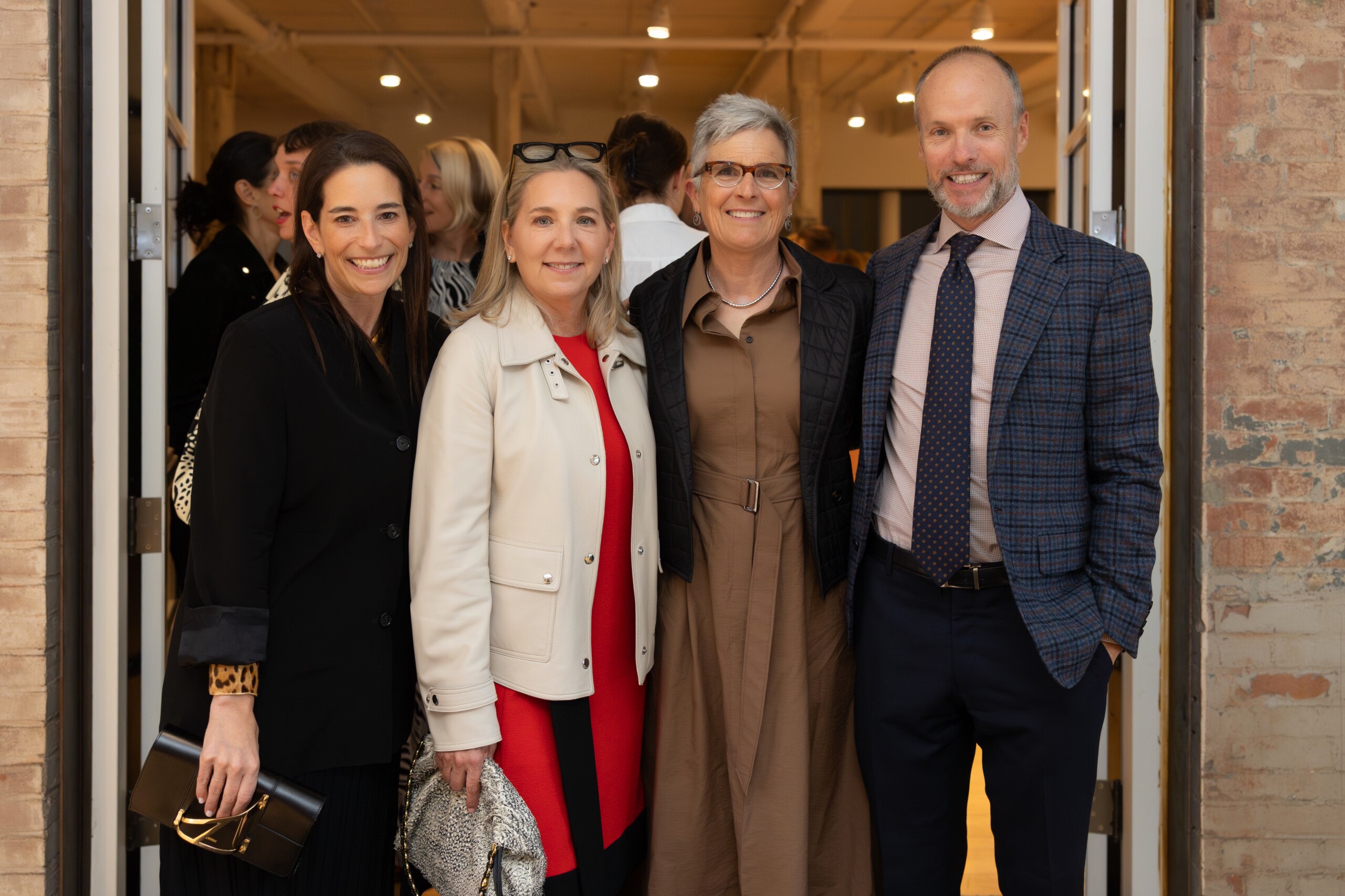 Four adults, three women and one man, stand closely together and smile for the camera at an indoor event. They are dressed in business or smart casual attire, with a brick wall and open doors behind them.