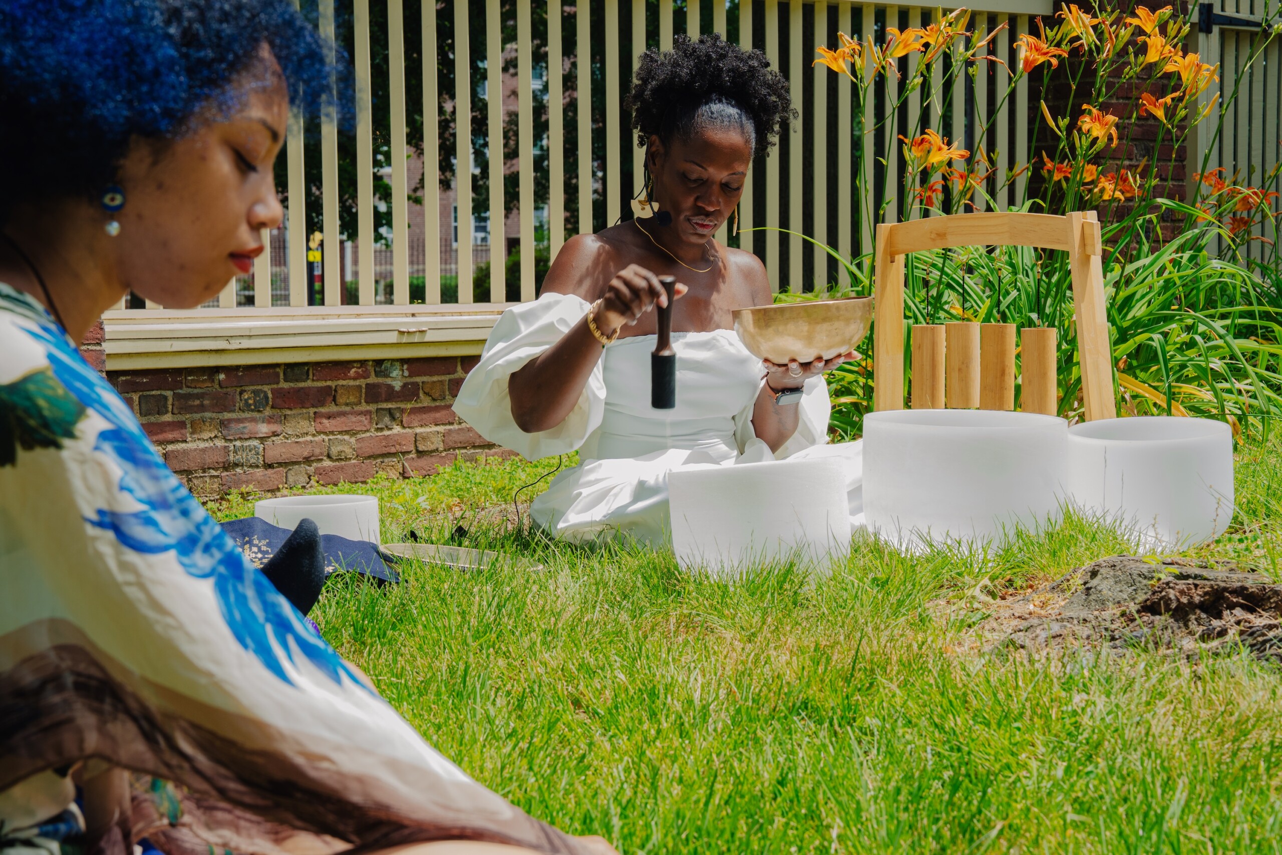 Two women sit on the grass outdoors. One woman in a white dress plays a singing bowl surrounded by white crystal bowls, while the other woman sits nearby with her eyes closed. Orange flowers and a fence are in the background.