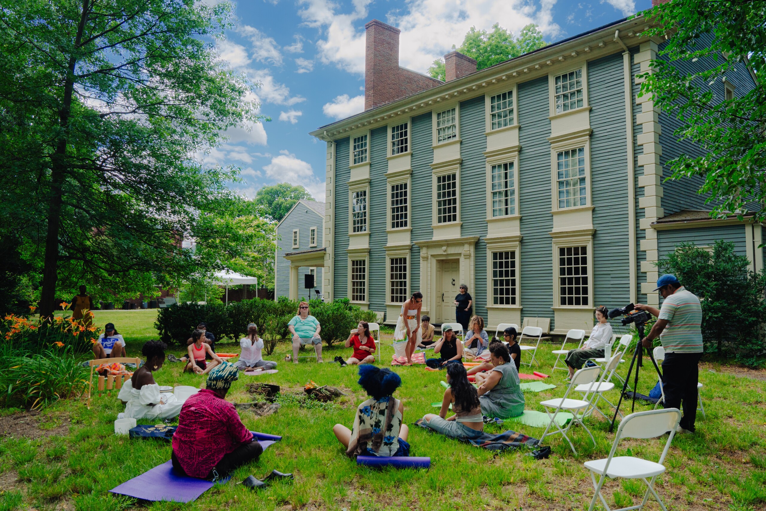 A group of people sit on yoga mats and chairs in a grassy area outside a large, historic house, surrounded by trees on a sunny day. Some are engaged in activities while others observe or film the event.