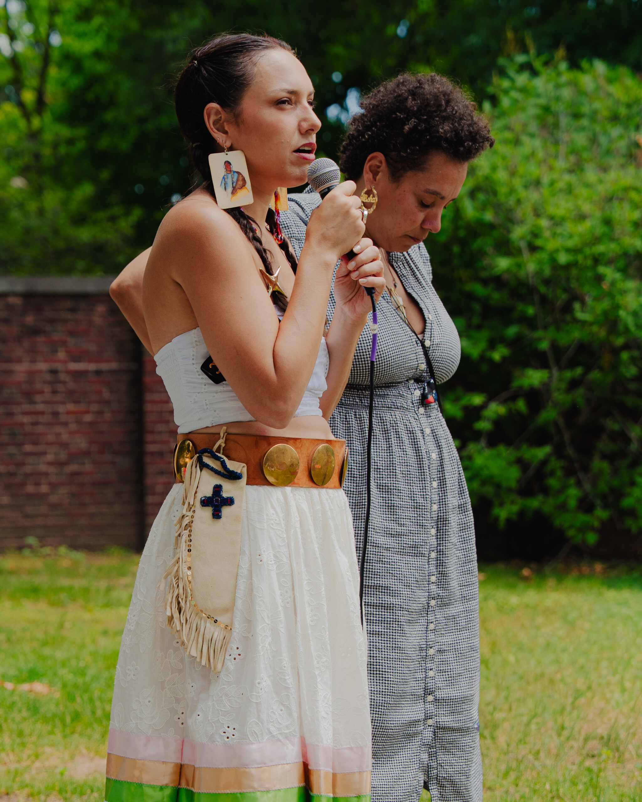 Two women stand outdoors; one speaks into a microphone wearing traditional attire with large earrings and a decorative belt, while the other stands beside her with eyes closed, both in a grassy area with trees and a brick wall in the background.