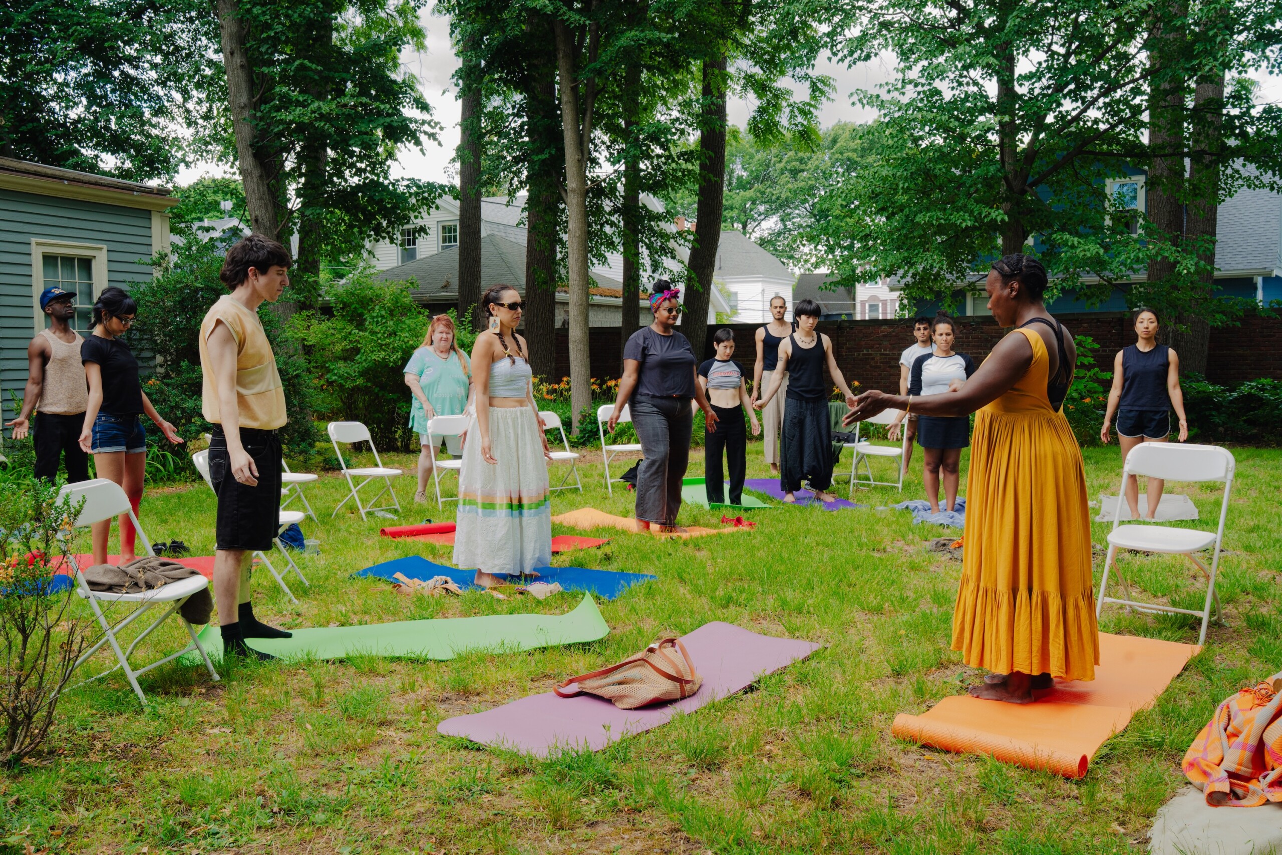 A group of people stand on colorful yoga mats in a circle on grass outdoors, with a woman in a yellow dress leading them. Folding chairs and trees surround the group in a residential backyard setting.